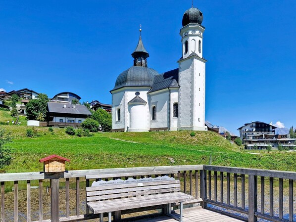 A church with two domes and a bell tower, surrounded by houses and a wooden deck with a bench.