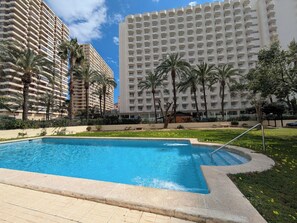 A swimming pool with a curved edge, surrounded by greenery and palm trees, with high-rise buildings in the background. - Waldorff by Interhome (Benidorm)