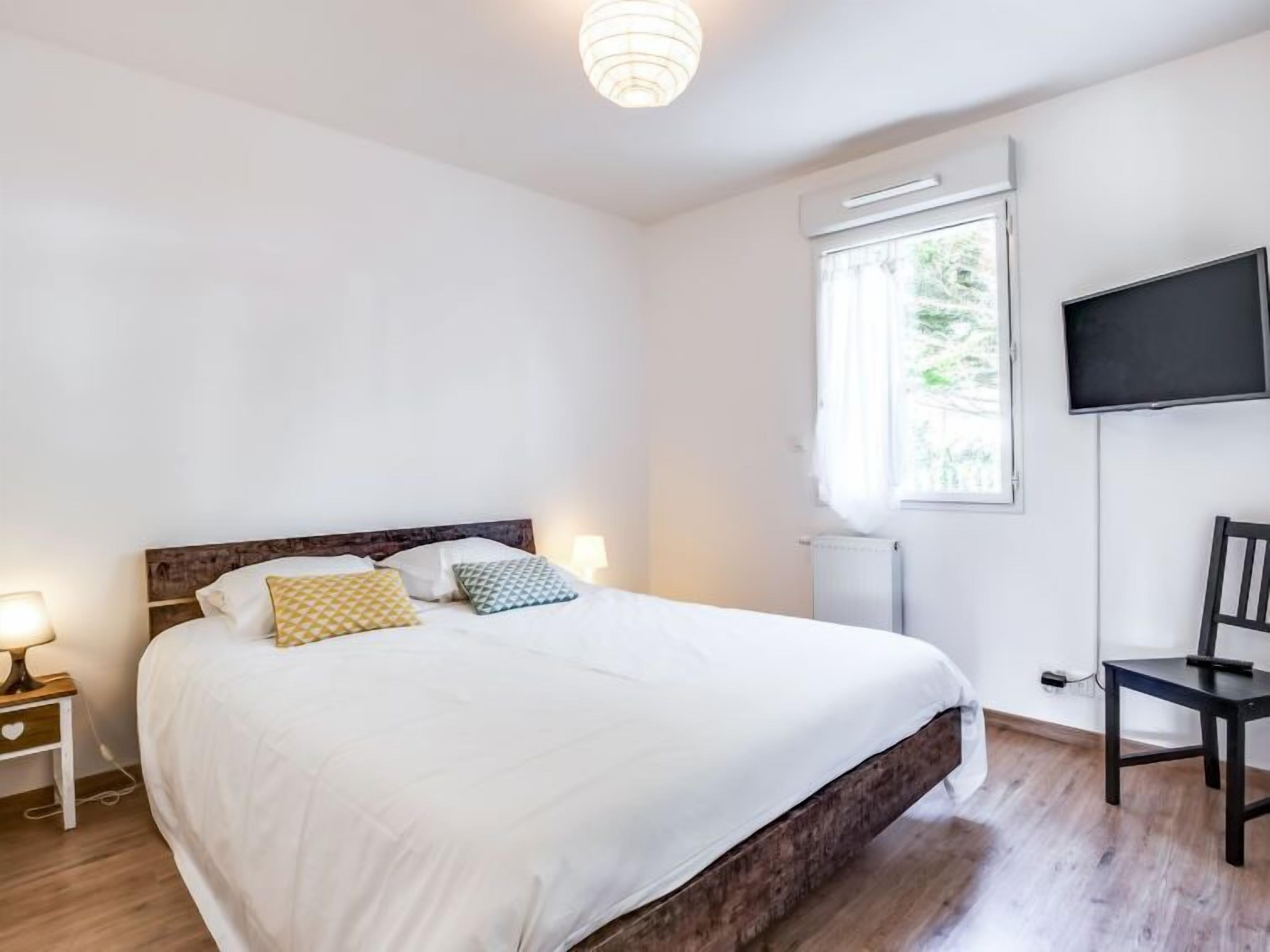 A bedroom with a large bed, a bedside table with a lamp, a chair, a TV mounted on the wall, and a window with curtains.