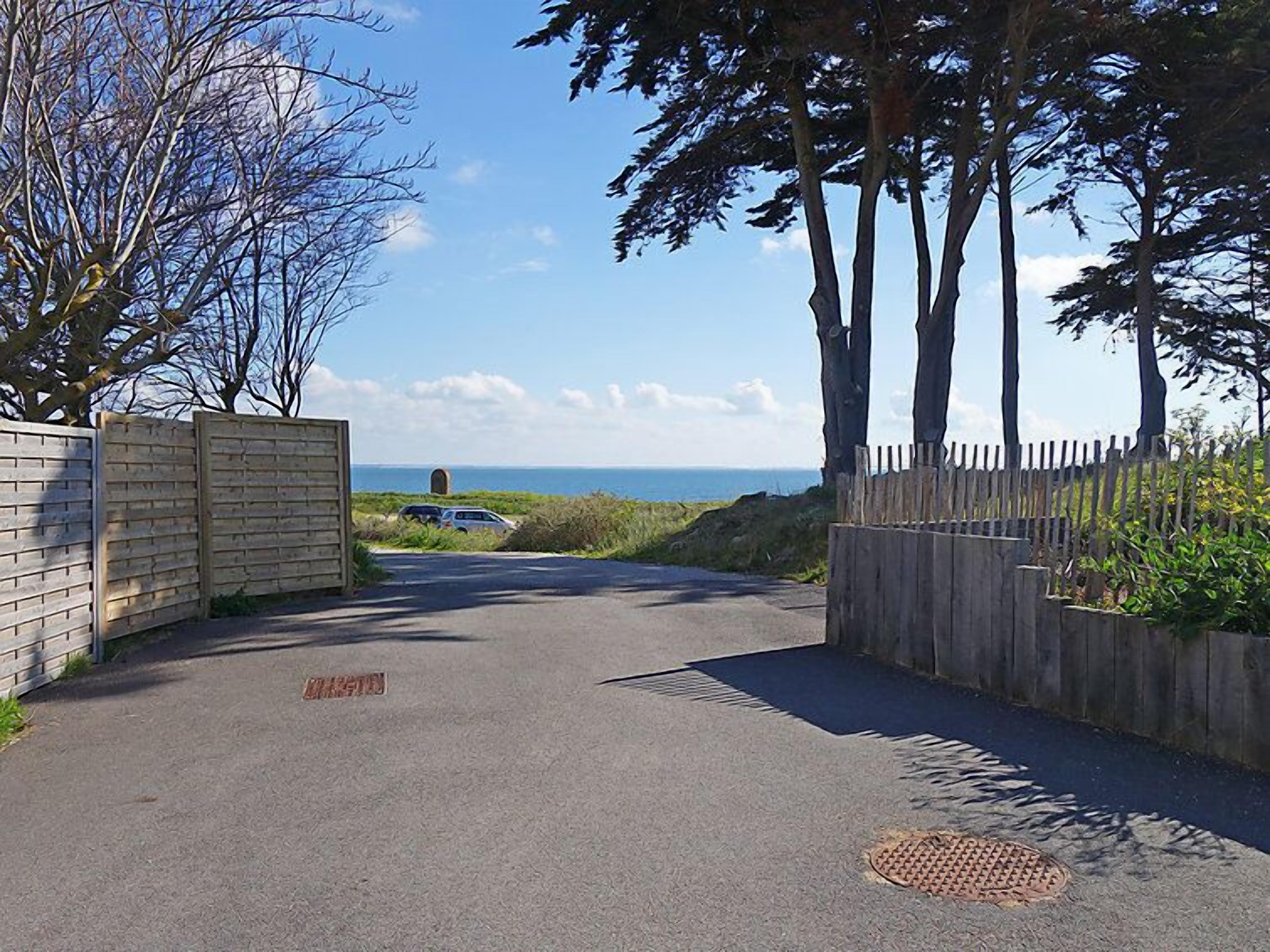 Una carretera pavimentada que conduce a una playa, con una cerca de madera a un lado y una reja metálica al otro.
