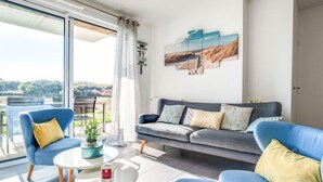 Un salon moderne avec un canapé gris, des fauteuils bleus et une table basse en verre. Une grande fenêtre offre une vue sur l’extérieur.