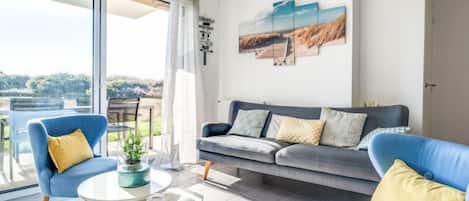 A modern living room with a grey sofa, blue armchairs, and a glass coffee table. There is a large window offering a view of the outdoors.