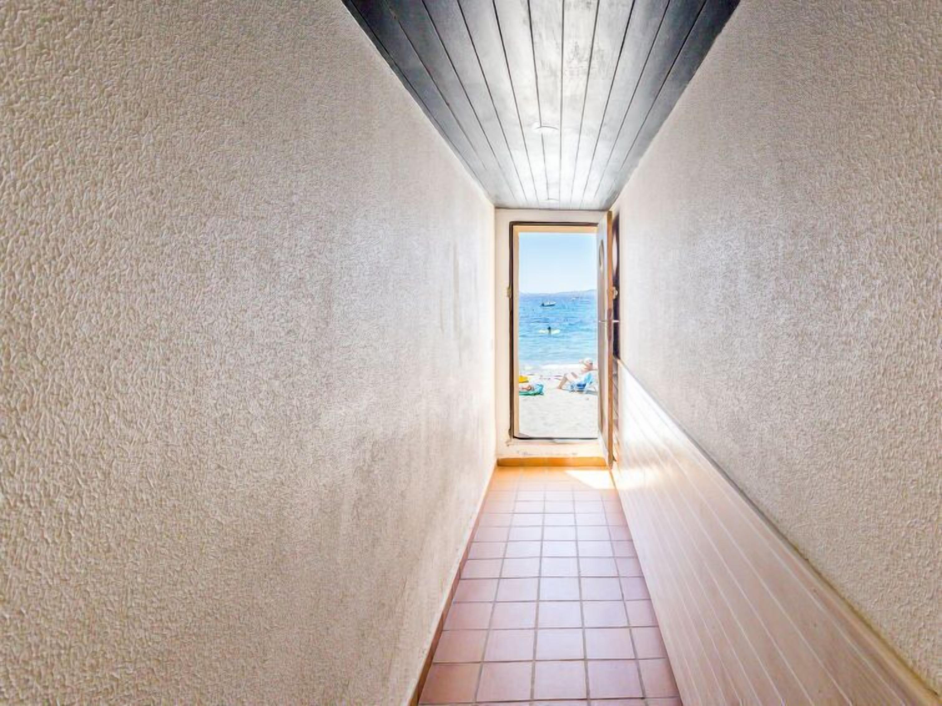 Un couloir étroit, dont le sol est recouvert de carreaux, mène à une porte qui donne sur une vue de la plage.