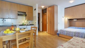 A compact kitchen with wooden cabinets, a dining table with chairs, and a bed with a blue blanket. - Apartment Apt 14 by Interhome (Canazei)