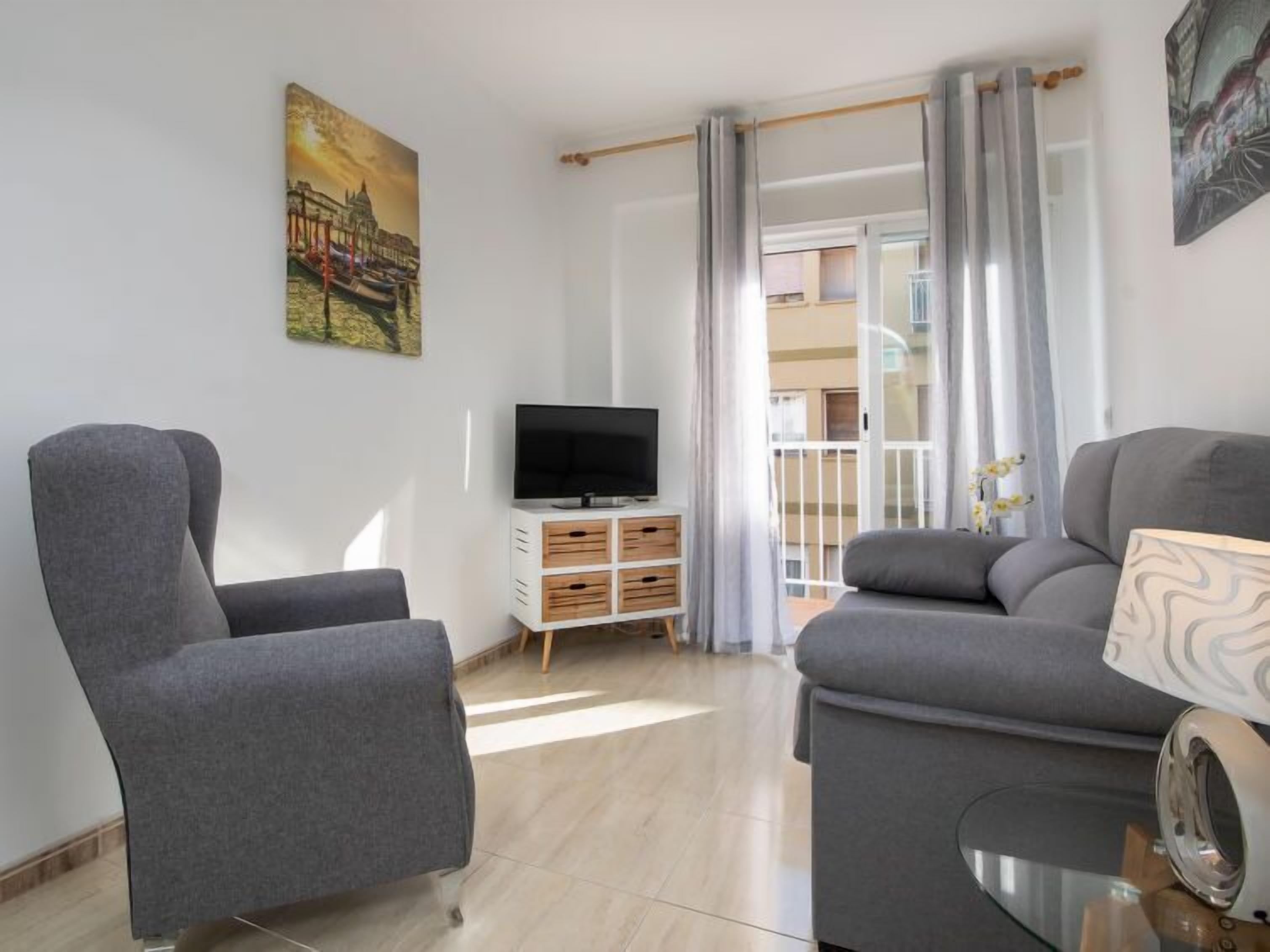 A modern living room with a grey sofa, a grey armchair, and a flat-screen TV on a wooden cabinet. There is a balcony with a view of the building and a painting of a cityscape on the wall.