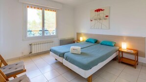A bedroom with two beds, a window with curtains, a radiator, a bedside table with a lamp, and a wall-mounted artwork. - Les Mimosas by Interhome (Saint Aygulf)