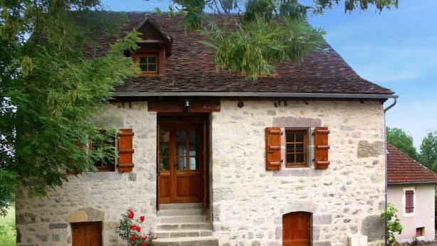 Ein Steinhäuschen mit einer Holztür, zwei Fenstern und Fensterläden sowie einer kleinen Veranda.
