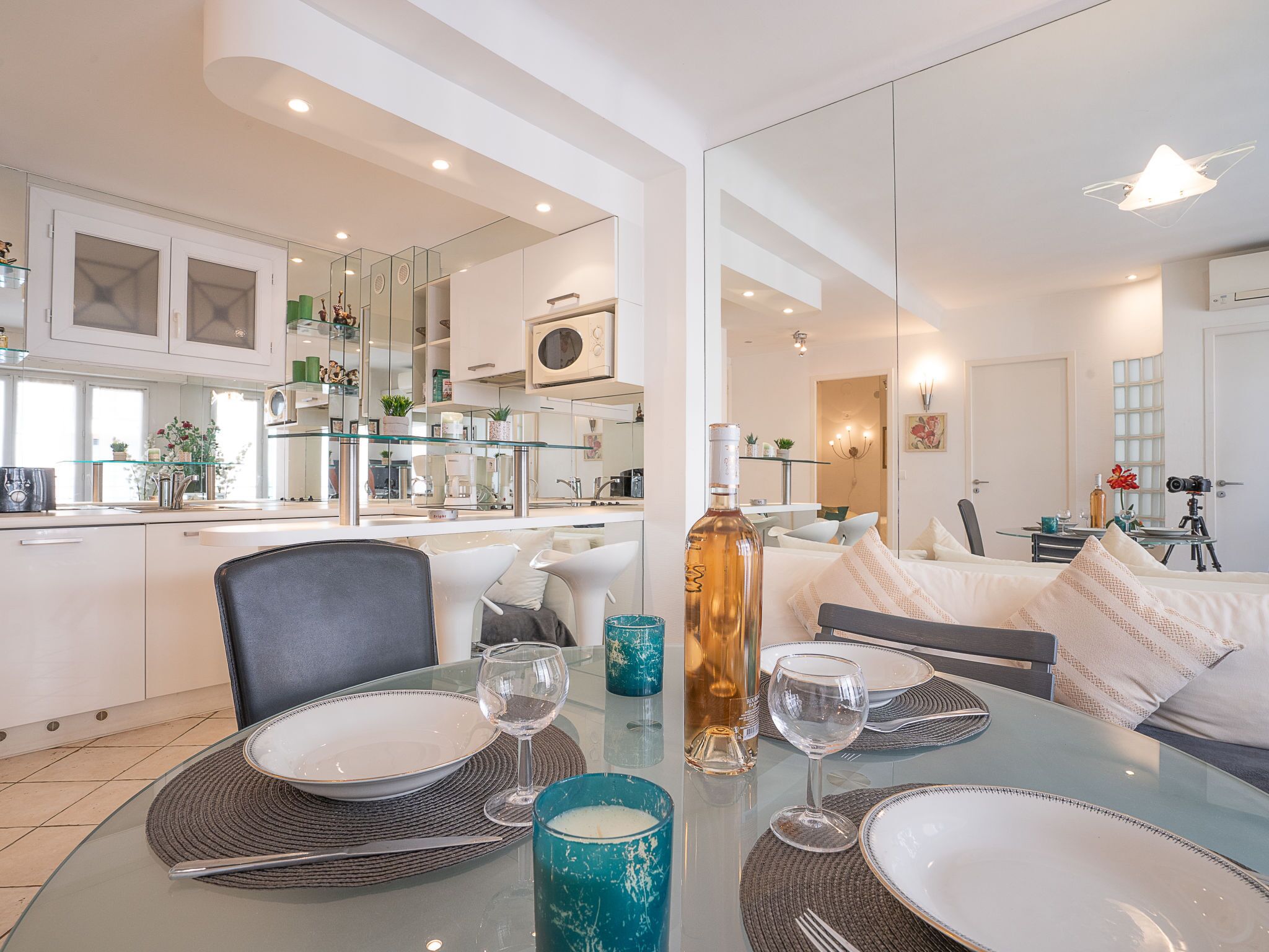 A modern dining area with a glass table set for four, surrounded by a kitchen and living space.