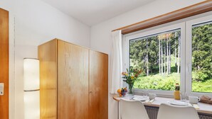 A modern kitchen with a wooden cabinet, white chairs, a dining table, and a window with a view of greenery. - Promenade Apt 216 by Interhome (Arosa)
