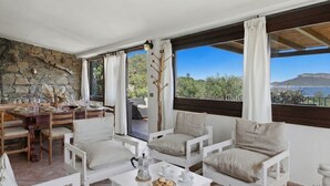 A room with a stone wall, a dining area with a table and chairs, a coffee table with a pot and pastries, and large windows with a view of trees and a body of water. - Vacation home Iride by Interhome (Golfo Aranci)