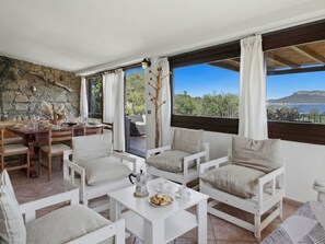 A room with a stone wall, a dining area with a table and chairs, a coffee table with a pot and pastries, and large windows with a view of trees and a body of water.