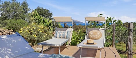 Two sun loungers with cushions, a book, and sunglasses on a table, surrounded by greenery and a view of the sea.
