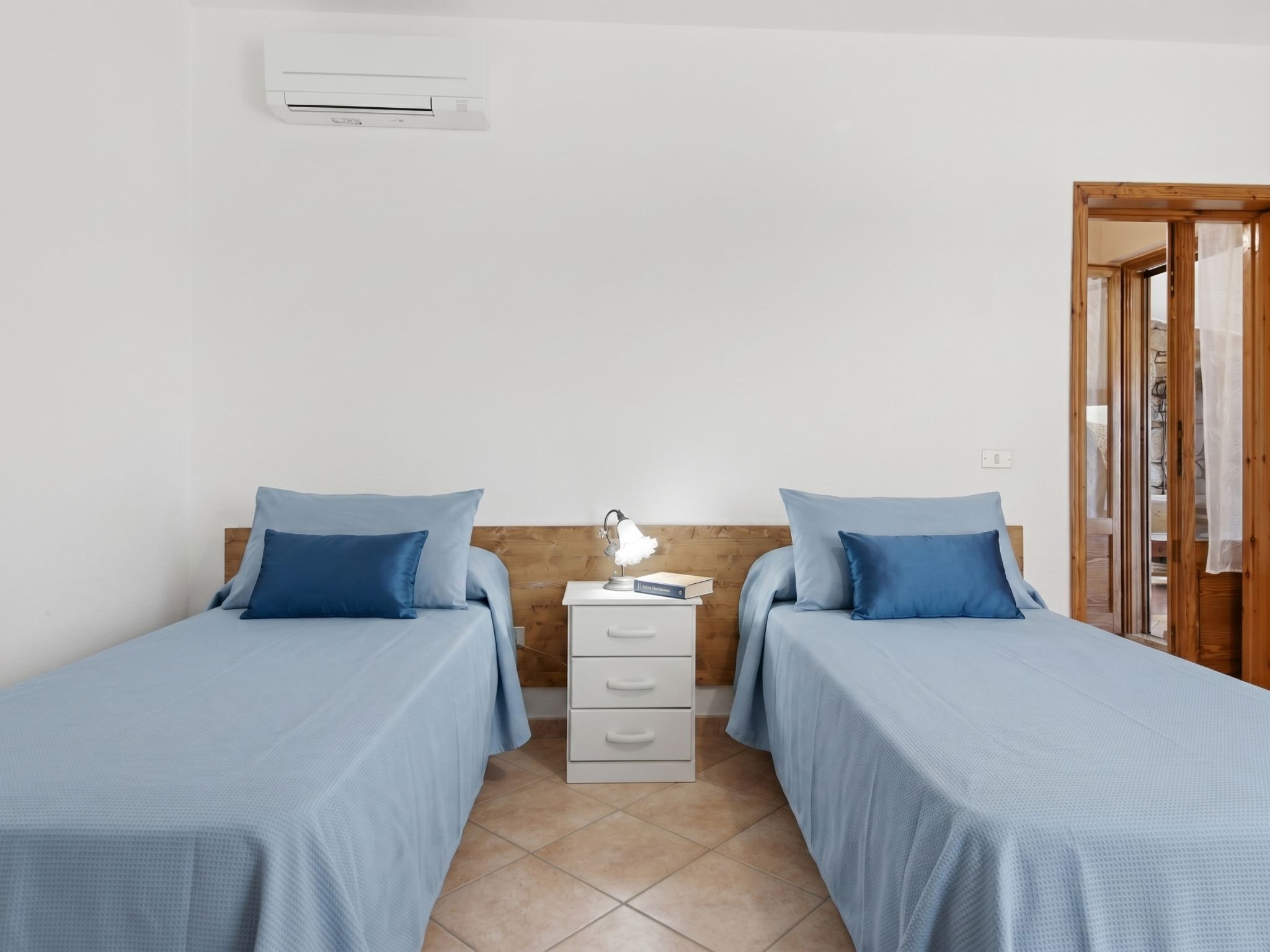 Una habitación con dos camas, una mesita de noche con lámpara, una pequeña cómoda y un equipo de aire acondicionado en la pared.