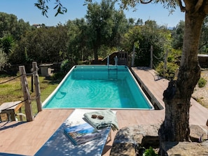A rectangular swimming pool with a wooden deck in the foreground, surrounded by trees and a stone wall.
