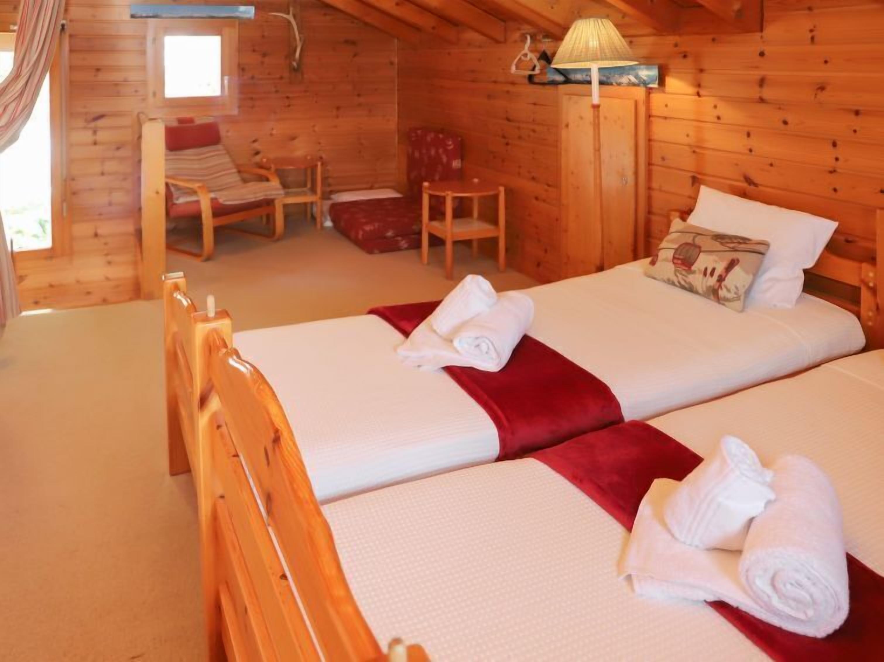 Quarto de cabana de madeira com duas camas, uma cadeira, uma mesa e um abajur.