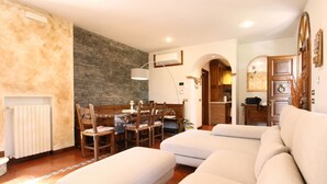 A living room with a stone wall, a dining area with wooden furniture, and a kitchen visible through an archway.