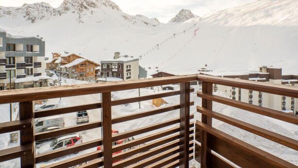 Sneet dækker bjergene i udsigten fra en balkon med trægelænder, med moderne bygninger og en stolelift i baggrunden.