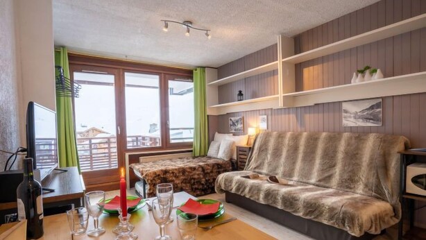 A cozy living room with a sofa, a dining table set with glasses and plates, a TV, and a window with curtains. - Le Curling B (Val Claret) by Interhome (Tignes)