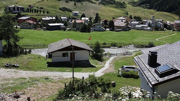 Uma aldeia rural com casas, um rio e montanhas ao fundo.