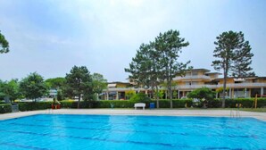 A rectangular swimming pool with clear blue water, surrounded by a tiled deck. In the background, there are multi-story buildings with balconies and a well-maintained lawn with trees. - Michelangelo by Interhome (Bibione)