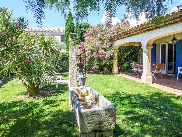 Um pátio com uma fonte de pedra, vegetação exuberante e uma área coberta com mesas e cadeiras ao ar livre.