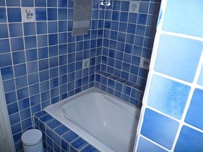 Casa de banho com azulejos azuis, uma banheira branca e um pequeno contentor de lixo.