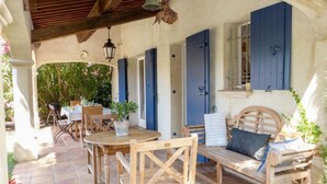 An outdoor patio with wooden furniture, a table, and chairs. There is a bench with cushions and a hanging bell. - Le Petit Olivier by Interhome (Les Issambres)