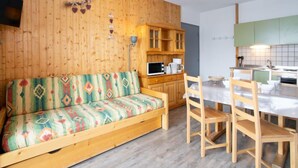 Un salon chaleureux avec un mur en bois, un canapé coloré, une table à manger avec des chaises et un coin cuisine doté de meubles verts.