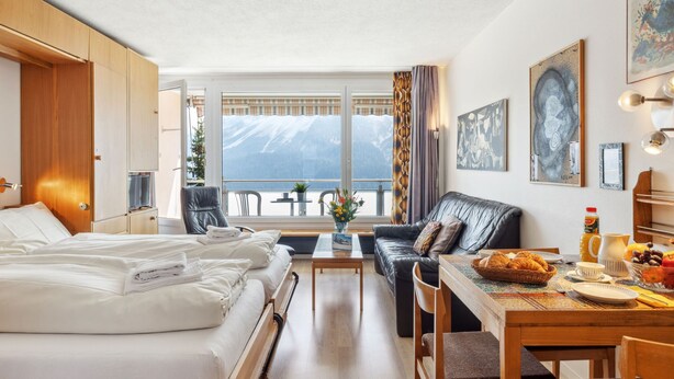 A hotel room with a bed, a sofa, a dining table with breakfast items, a window with a mountain view, and artwork on the walls. - Promenade Apt 210 by Interhome (Arosa)
