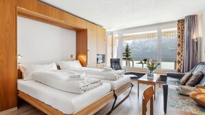 A hotel room with a bed, a small table, and a view of mountains through large windows. - Promenade Apt 210 by Interhome (Arosa)