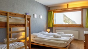 Une chambre avec un lit superposé, un lit simple, une fenêtre avec des rideaux, un radiateur et une commode en bois.