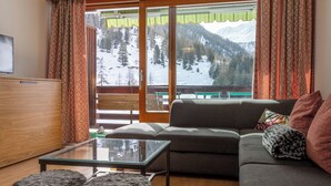 Un salon moderne avec un canapé, des poufs et une table basse en verre. Un balcon avec vue sur des montagnes enneigées est visible à travers les portes vitrées.