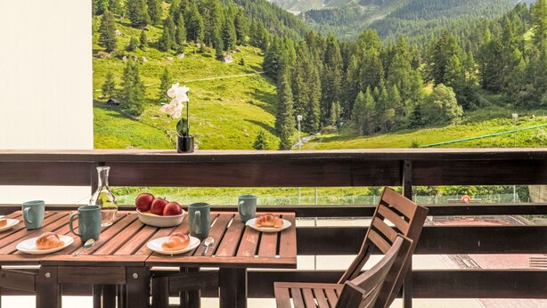 Ein Balkon mit einem Frühstückstisch, mit Blick auf eine gebirgige Landschaft unter blauem Himmel.