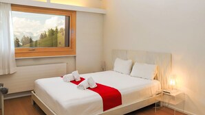 Ein ordentlich bezogenes Bett mit weißen Leinen und einem roten Akzent, ein Nachttisch mit Lampe sowie ein Fenster mit Blick auf eine grüne Landschaft.