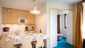 Eine kompakte Küche mit Mikrowelle, Herd und Kaffeemaschine. Im angrenzenden Raum befindet sich ein Esstisch mit Stühlen sowie ein kleines Bett.
