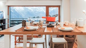 Ein Essbereich mit Tisch für vier Personen, Blick auf schneebedeckte Berge und eine Küchenzeile mit Herd und Kochgeschirr.