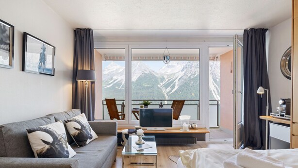 A modern hotel room with a sofa, a bed, a desk with a computer, and a view of snow-capped mountains. - Promenade Apt 409 by Interhome (Arosa)