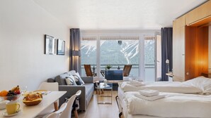 A hotel room with a bed, a sofa, a dining table with breakfast, a balcony with a mountain view, and a desk with a lamp. - Promenade Apt 409 by Interhome (Arosa)