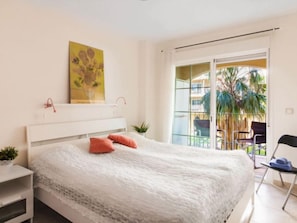 A modern bedroom with a large bed, a small bedside table, a potted plant, and a balcony with a view of palm trees.