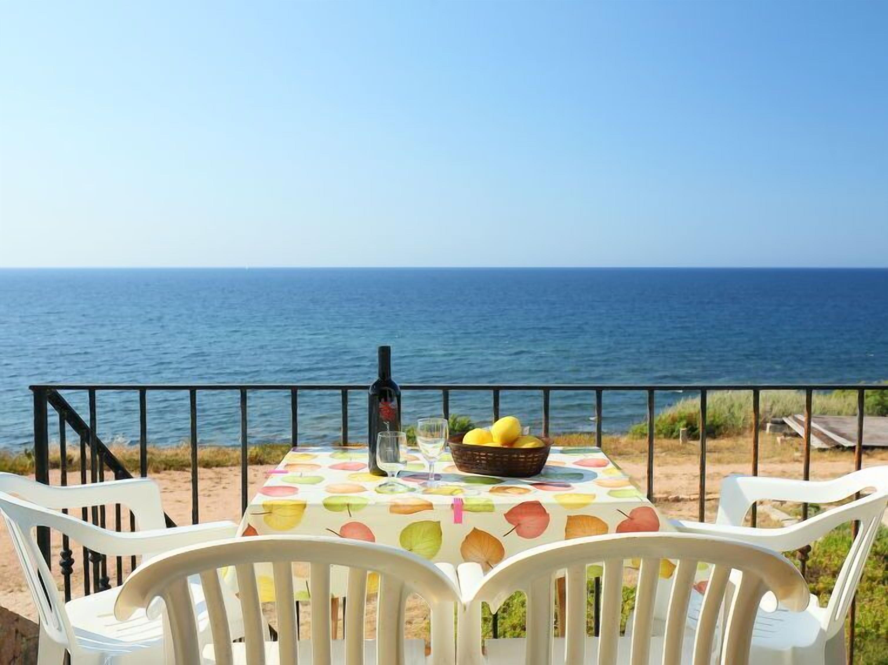 Une table recouverte d’une nappe à motifs de fruits, une bouteille et un saladier rempli de fruits, le tout placé devant une vue sur la mer.