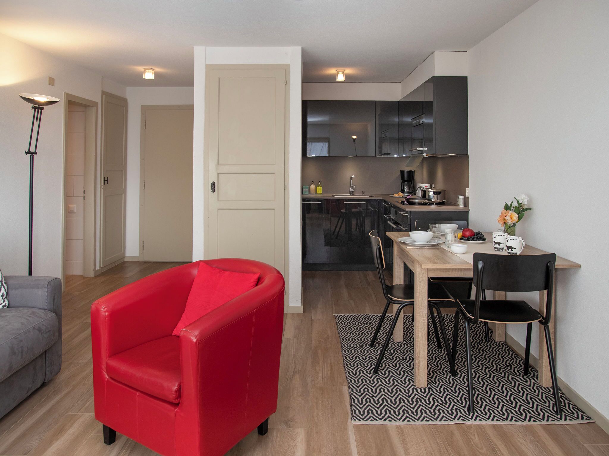 A modern apartment with a dining area, a red chair, a grey sofa, and a kitchen in the background.