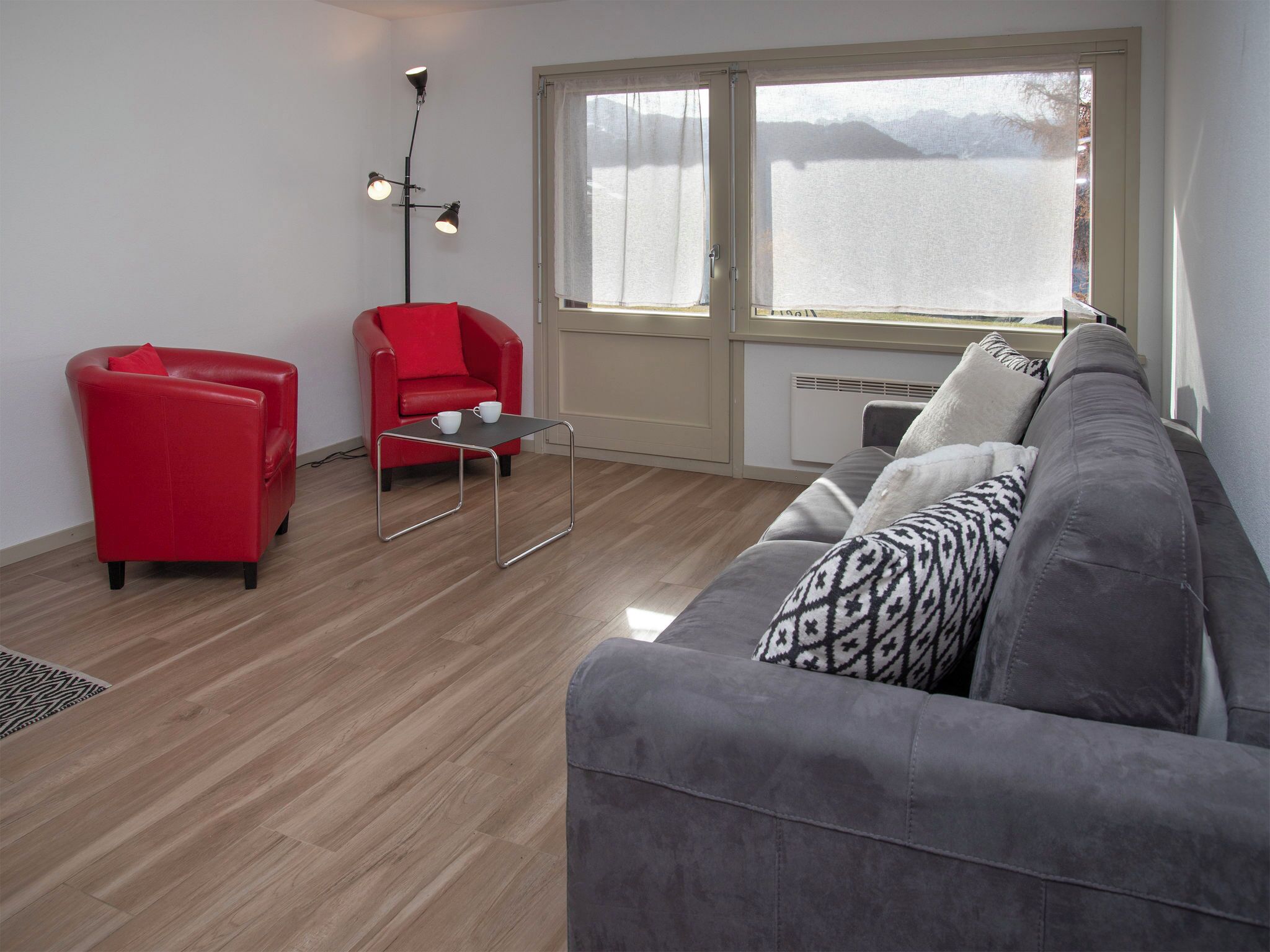 Ein modernes Wohnzimmer mit einem roten Sessel, einem grauen Sofa und einem kleinen Tisch mit zwei weißen Tassen.