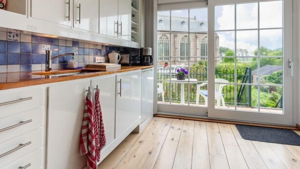 A modern kitchen with white cabinets, a wooden countertop, and a view of a large house through a sliding glass door.