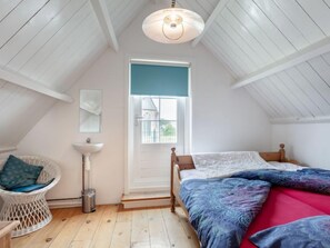 A cozy attic bedroom with a sloped ceiling, a bed with patterned bedding, a white wicker chair, a bedside table, and a window with a view.