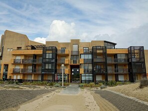 En moderne, flereetagers boligbygning med altaner, en central indgang og en belagt sti.