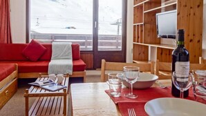 A cozy room with a red sofa, wooden coffee table, and a dining table set for two. A television is mounted on the wall, and a bottle of wine is on the table. - Les Tommeuses (Val Claret) by Interhome (Tignes)