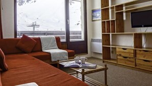 A cozy living room with a sofa, a coffee table with books and cups, a TV on a wooden shelf, and a large window with a snowy view. - Les Tommeuses (Val Claret) by Interhome (Tignes)