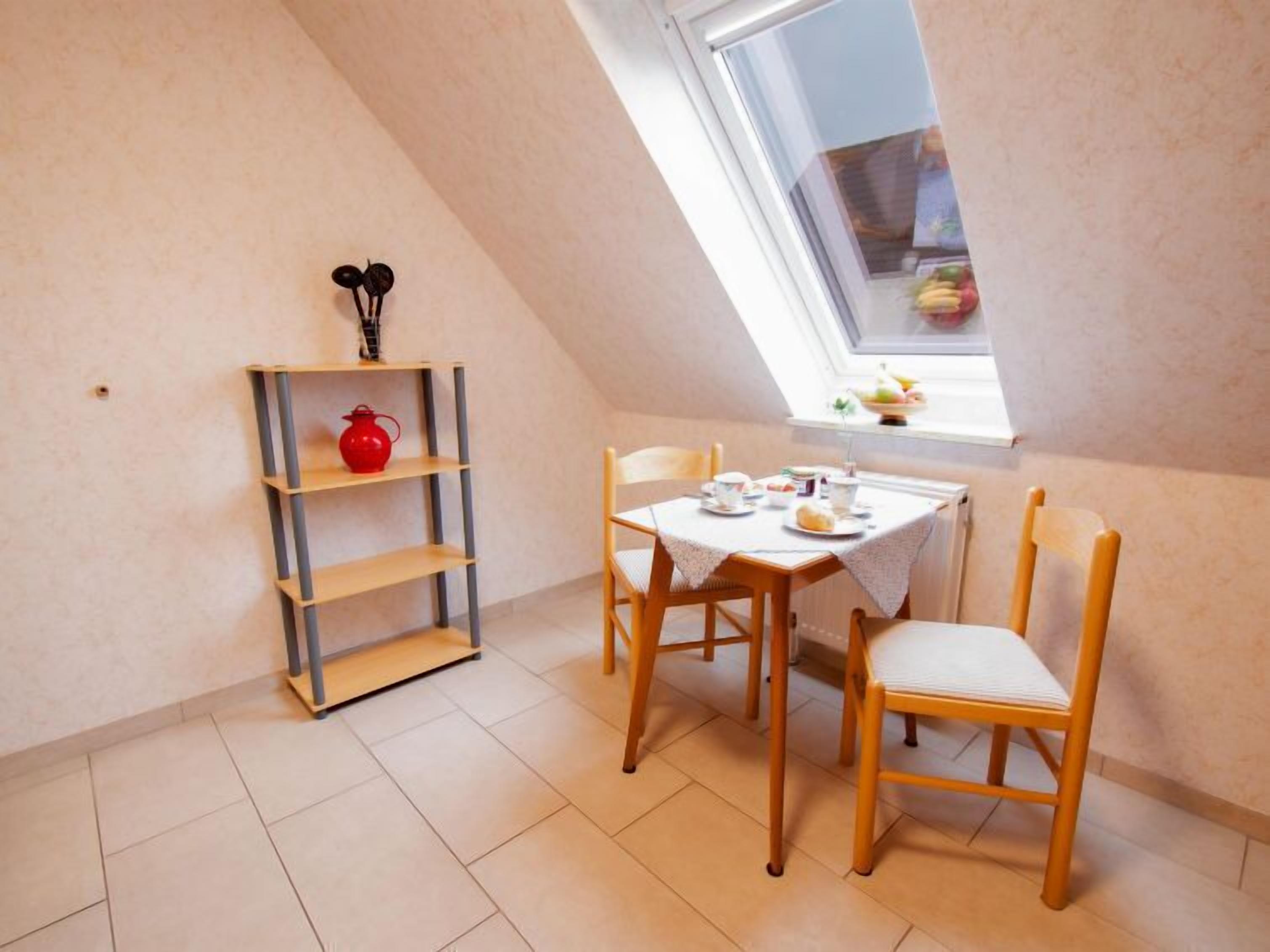 Ett litet matområde med ett bord och stolar, en röd tekanna på en hylla och ett fönster med utsikt över fruktträd.