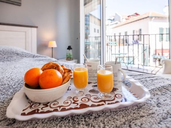 A breakfast tray with oranges, croissants, and orange juice on a bed with a view of buildings outside.