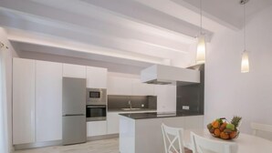 Uma cozinha moderna com armários brancos, eletrodomésticos de aço inoxidável e uma ilha central. Há uma mesa de jantar com cadeiras e uma fruteira na ilha.